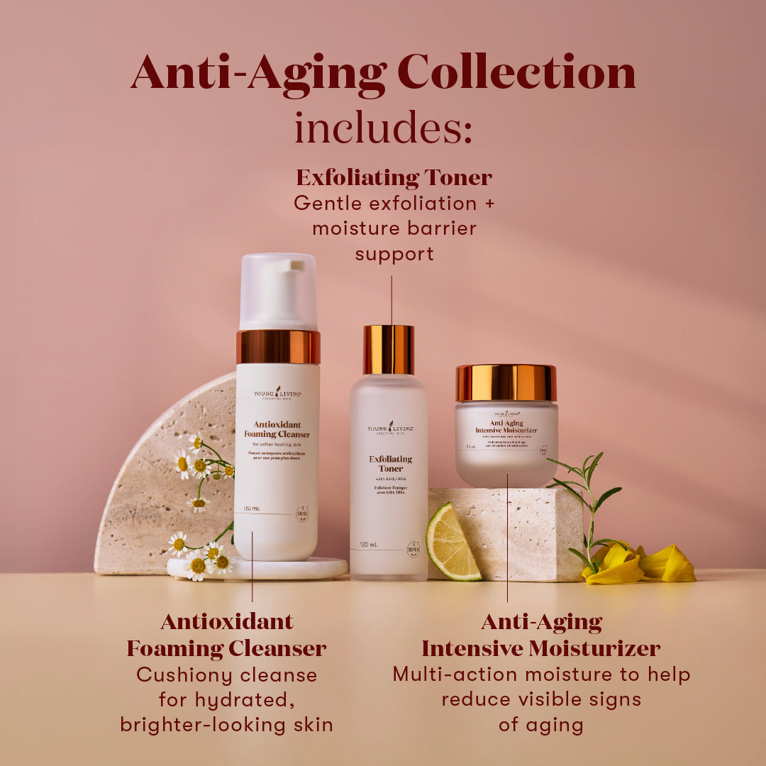 Three skincare products arranged on a beige surface against a dusty pink background, with text labels describing the Anti-Aging Collection:  Antioxidant Foaming Cleanser, Exfoliating Toner, and Anti-Aging Intensive Moisturizer. Decorative stones, chamomile flowers, and a lime wedge are placed around the products.