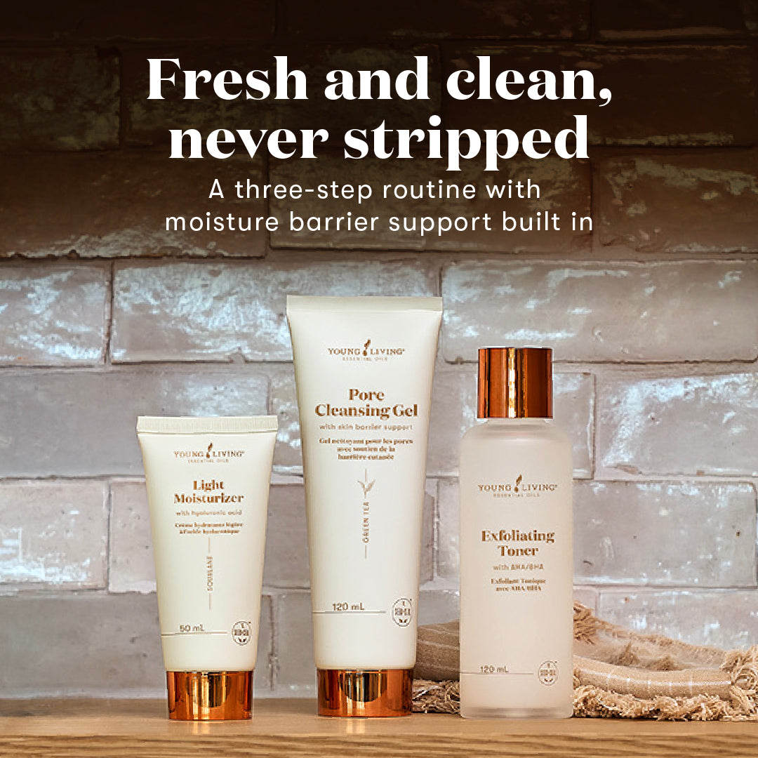 Three skincare products from Young Living are displayed on a wooden surface against a rustic gray brick wall background. The products are a Light Moisturizer tube, a Pore Cleansing Gel tube, and an Exfoliating Toner bottle, all in white with metallic copper accents.