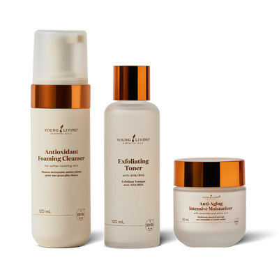 Three skincare products from Young Living on a white background: a frosted bottle of Antioxidant Foaming Cleanser, a frosted bottle of Exfoliating Toner with a metallic copper cap, and a frosted jar of Anti-Aging Intensive Moisturizer with a metallic copper lid.