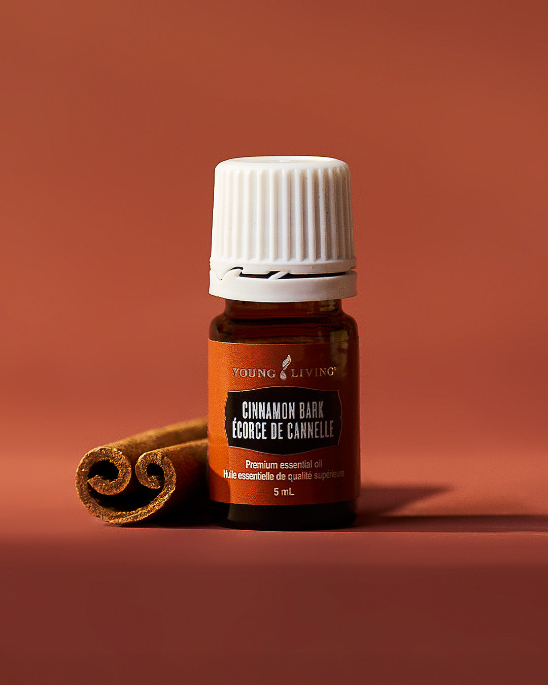 A 5 mL bottle of Young Living Cinnamon Bark essential oil with a white ribbed cap and an amber-brown label, styled with a cinnamon stick on a warm terracotta background.