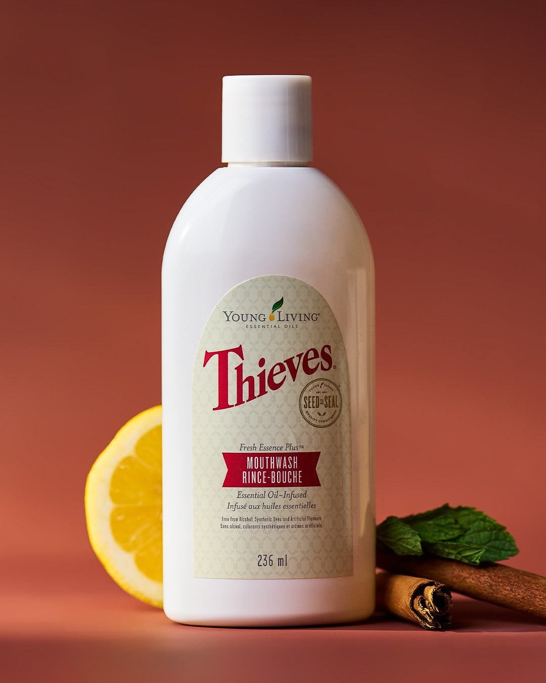 A bottle of Young Living Thieves Mouthwash Rinse essential oil blend, styled with a sliced orange, mint leaves, and cinnamon sticks on a warm terracotta background.