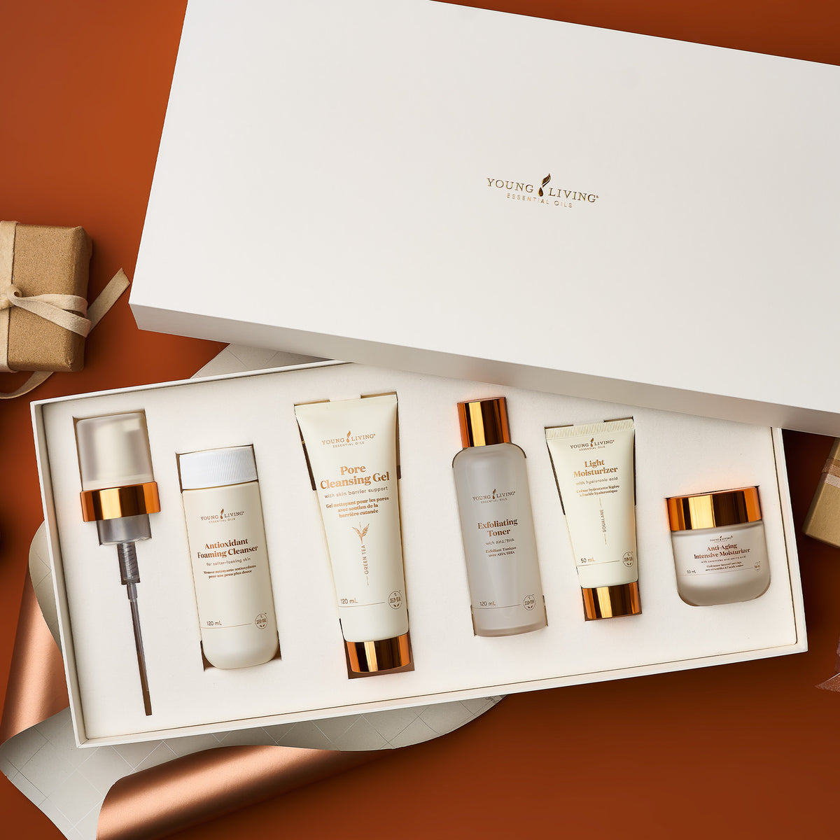 A Young Living Skincare gift set displayed in an open white box on a warm brown background. The set includes a cleansing brush, cleansing gel, toner, body wash, body lotion, and a cream jar, all in elegant white packaging with metallic gold caps and lettering.