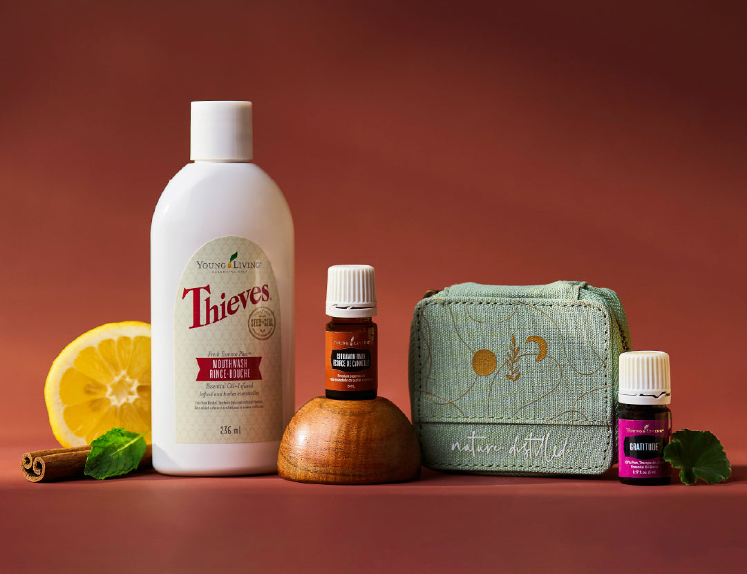 A curated wellness gift set featuring a large bottle of Thieves mouthwash, a small bottle of Young Living essential oil, a mint green fabric pouch with gold embroidery, and a small essential oil bottle, styled with a lemon slice, cinnamon sticks, and mint leaves on a warm terracotta background.