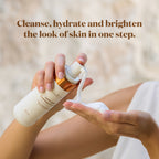 A person holds a white skincare pump bottle with a gold collar and dispenses white cream into their hand. Brown text at the top reads, 'Cleanse, hydrate and brighten the look of skin in one step.