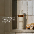 A bottle of Antioxidant Foaming Cleanser stands on a tiled shelf next to a wooden brush, with text on the left reading 'Clearer, brighter-looking skin with every wash.'.