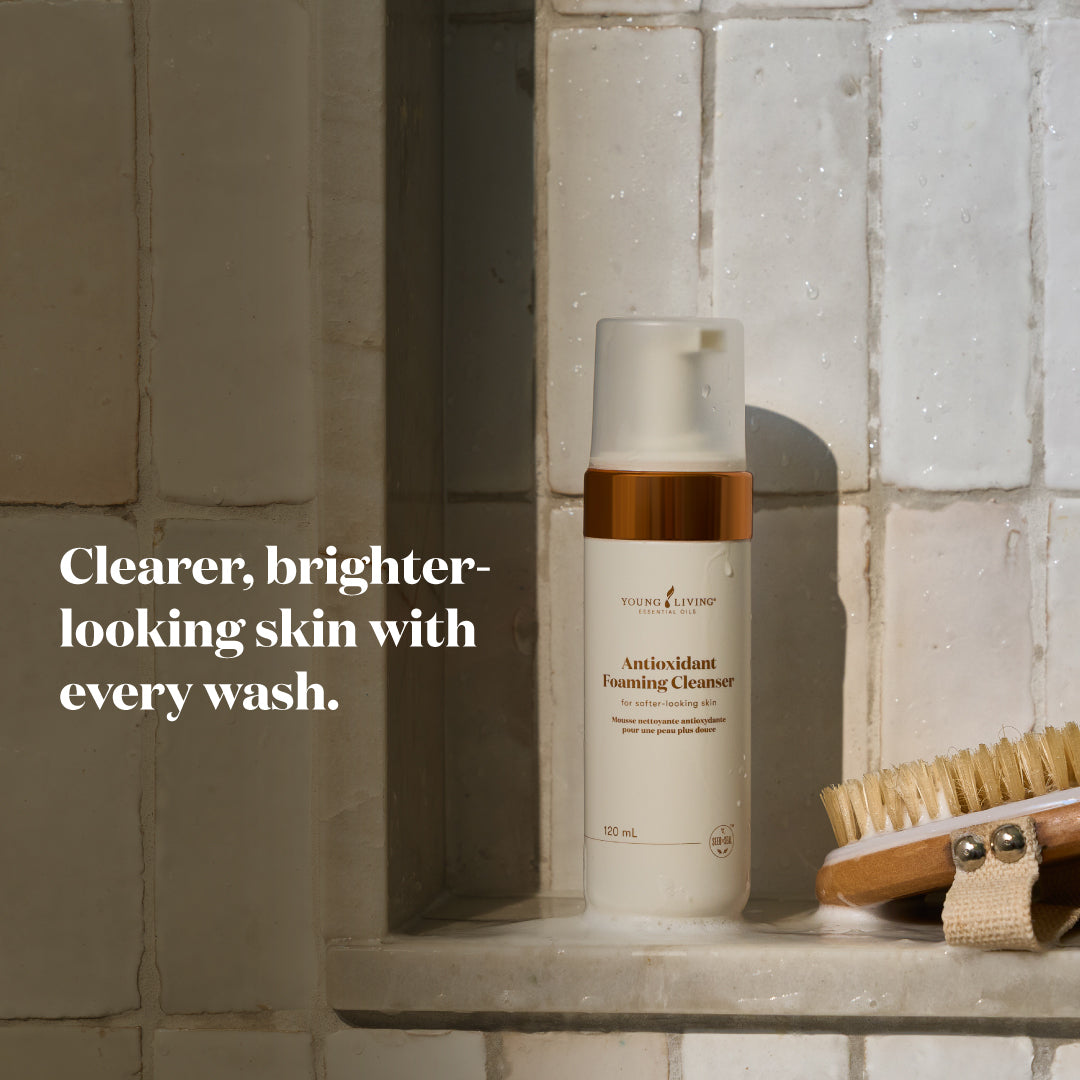 A bottle of Antioxidant Foaming Cleanser stands on a tiled shelf next to a wooden brush, with text on the left reading 'Clearer, brighter-looking skin with every wash.'.