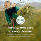 A person in a straw hat with colorful pom-poms, a green long-sleeve top, and a face covering bends over in a field while holding a large bundle of dried yellow flowers. White text over the photo reads, 'Farm-grown care in every cleanse.