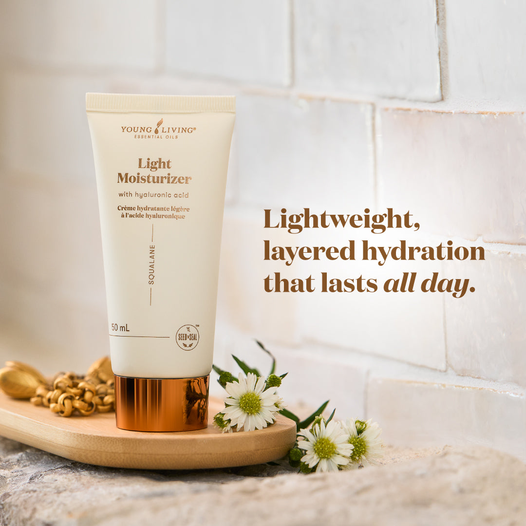 A 50 mL tube of Young Living Light Moisturizer with hyaluronic acid, featuring a cream-colored body and a metallic copper cap, displayed on a wooden tray with daisies and walnuts against a white brick wall background.