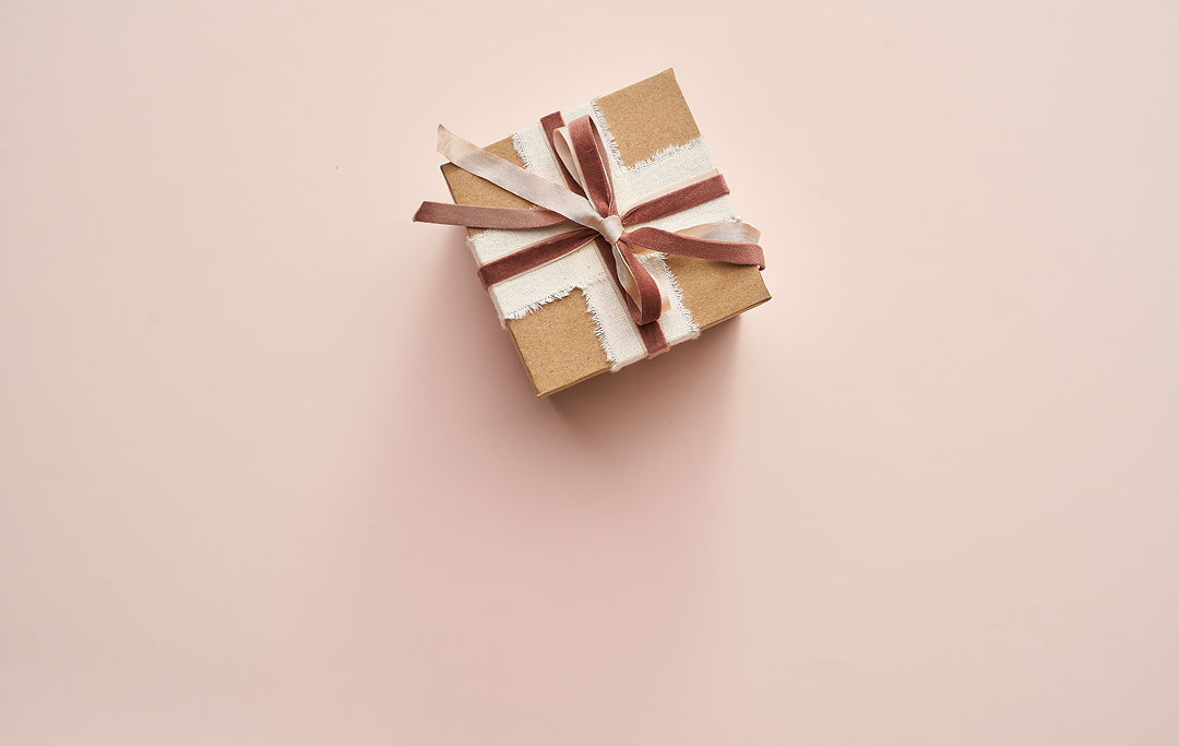 A small square gift box wrapped in brown kraft paper with a white paper band and tied with a brown ribbon, placed on a solid pale pink background.