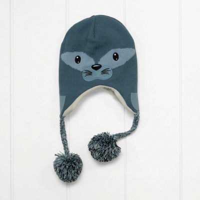 A dark teal knit winter hat with a seal face design on the front, featuring ear flaps with a light gray lining and two braided tassels ending in pom-poms, shown against a white paneled background.